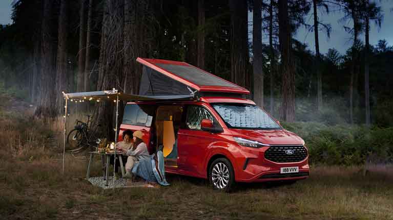 Red Ford Tourneo Connect campervan with open roof, parked in woodland. Two people relax at a table under a lit awning