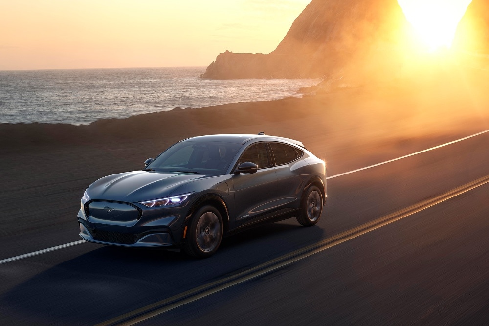A Ford Mustang Mach-E electric car drives along a coastal road at sunset.