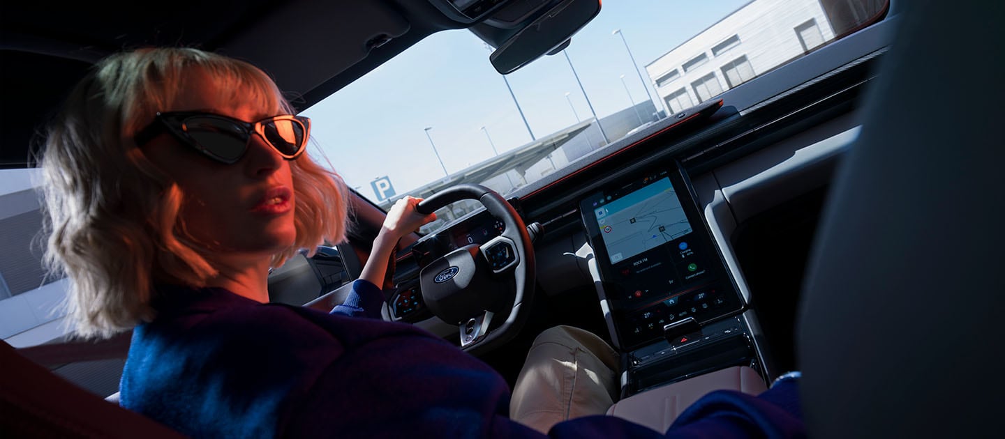 A woman sits in the driving seat of the electric Ford Capri, also showing the large 14.6" SYNC Move touchscreen.
