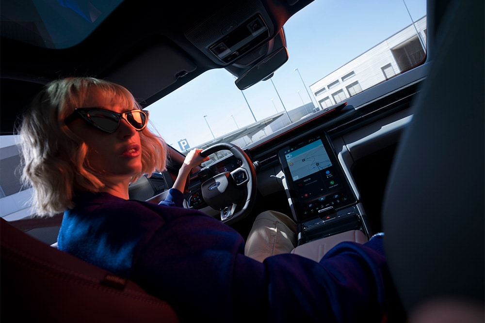 A woman sits in the driving seat of the electric Ford Capri, also showing the large 14.6" SYNC Move touchscreen.