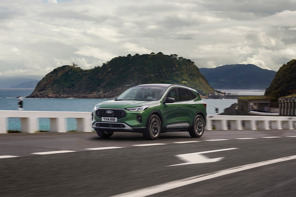 A green Ford Kuga self-charging hybrid car driving along a scenic road. 