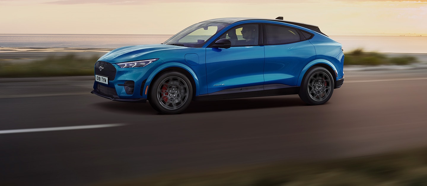 A blue electric Ford Mustang Mach-E driving along a coastal road at sunset.