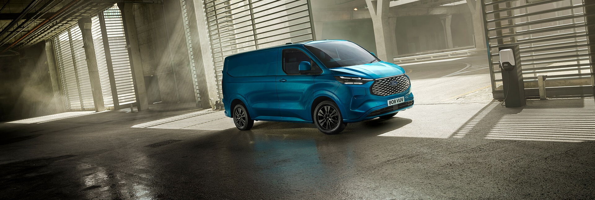 A blue Ford E-Transit Custom electric van driving through a carpark.