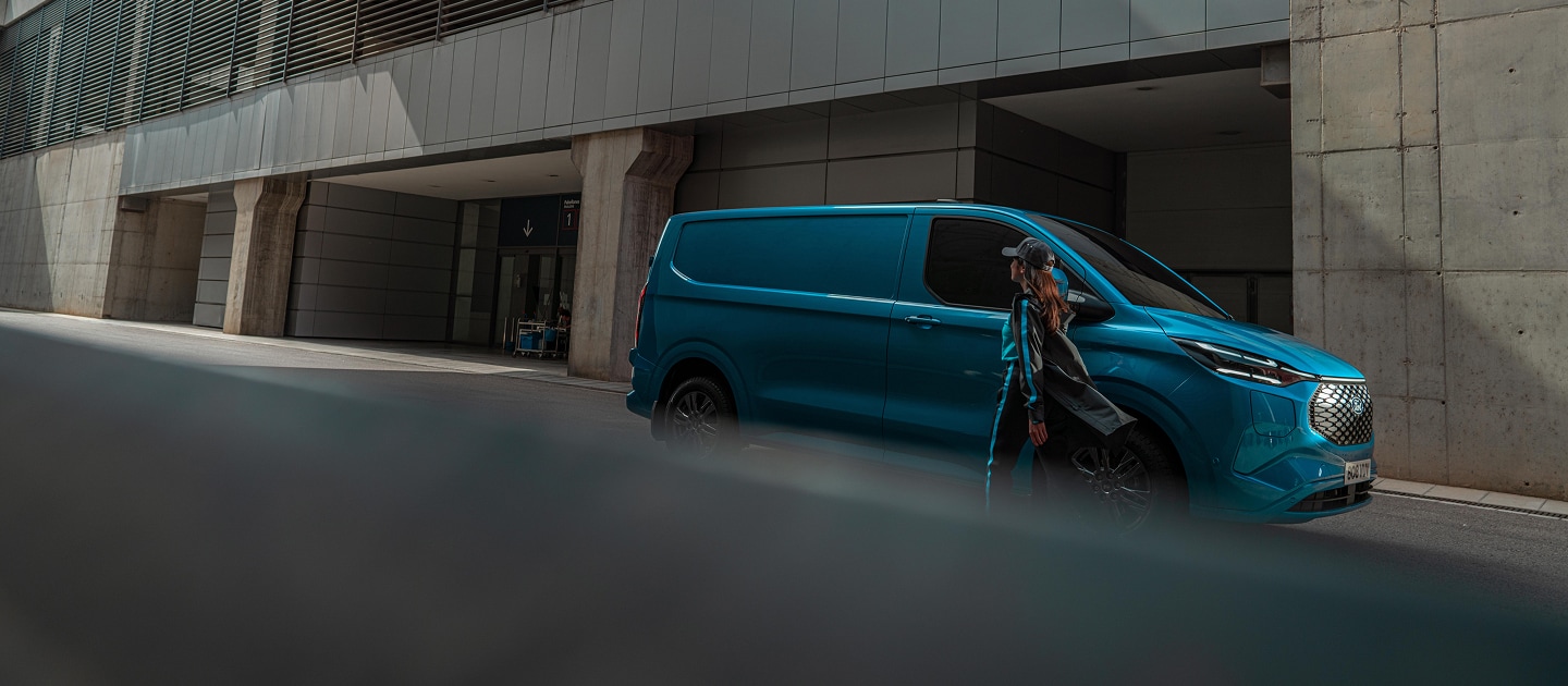 A woman walks past a blue Ford E-Transit Custom electric van parked on a street.
