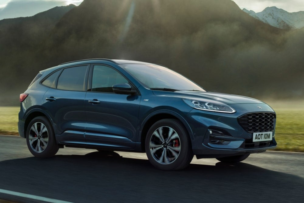 A blue Ford Kuga SUV driving on a winding road with mountains in the background.