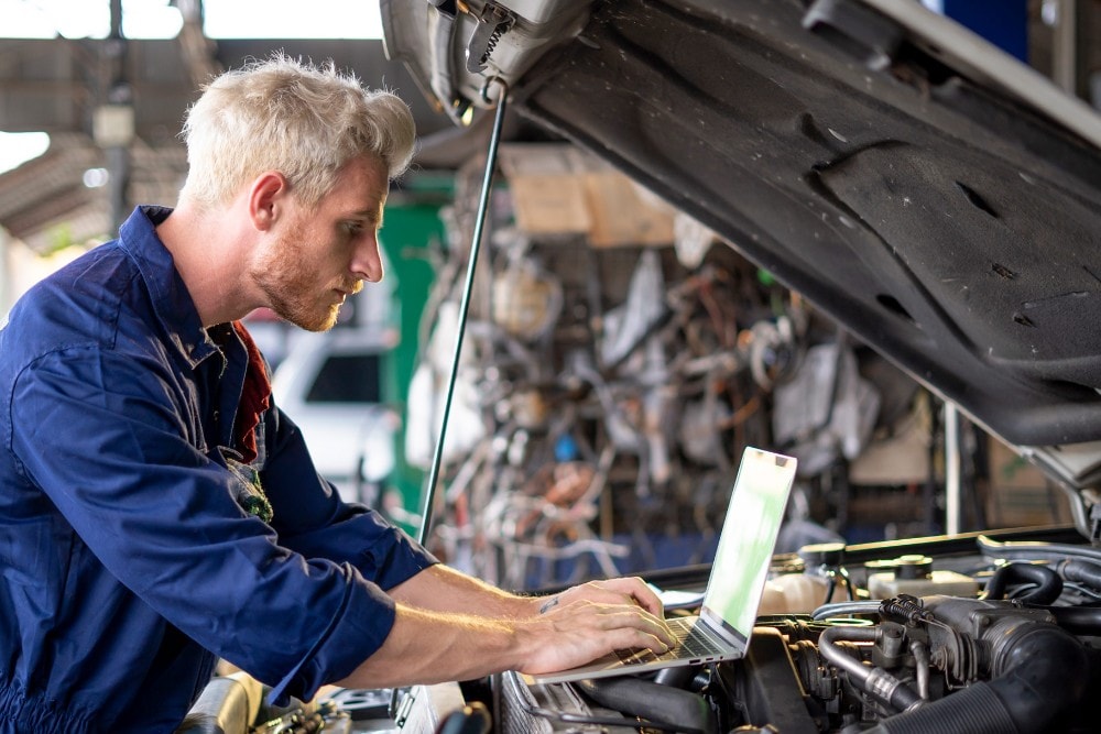 A technician uses a laptop while working on the engine of a vehicle with the bonnet open.