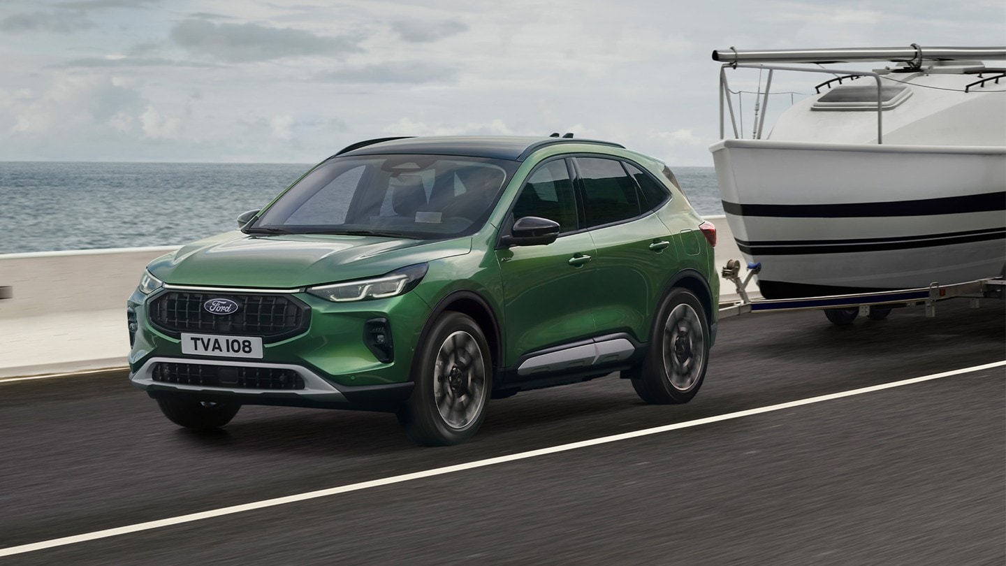 A green Ford Kuga towing a small boat along a coastal road.