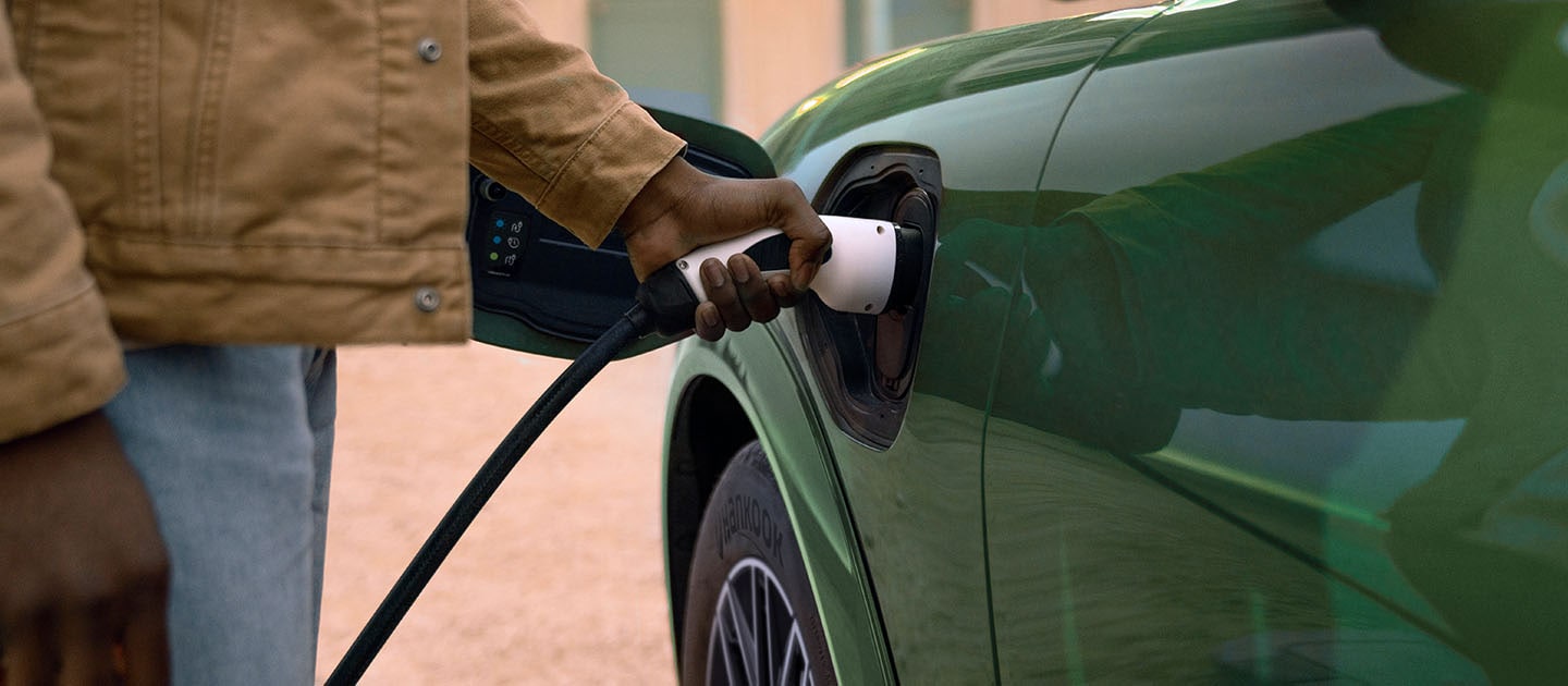 A person in a cream jacket plugging an electric charging cable into their green electric Ford Mustang Mach-E.