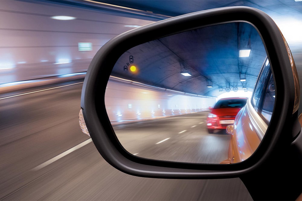 A close-up of the side-view mirror in a tunnel, showing a red car approaching in the driver's blind spot.