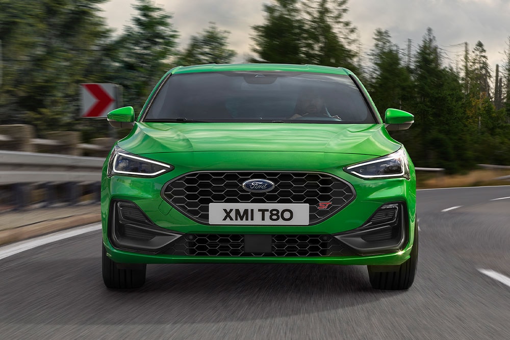 The front of a Mean Green Ford Focus ST going around a corner, with blurred trees in the background.