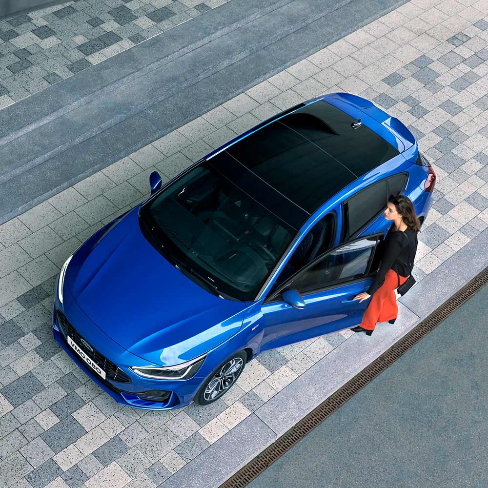 An overhead view of a woman getting into her blue Ford Focus that has a glass panoramic roof.
