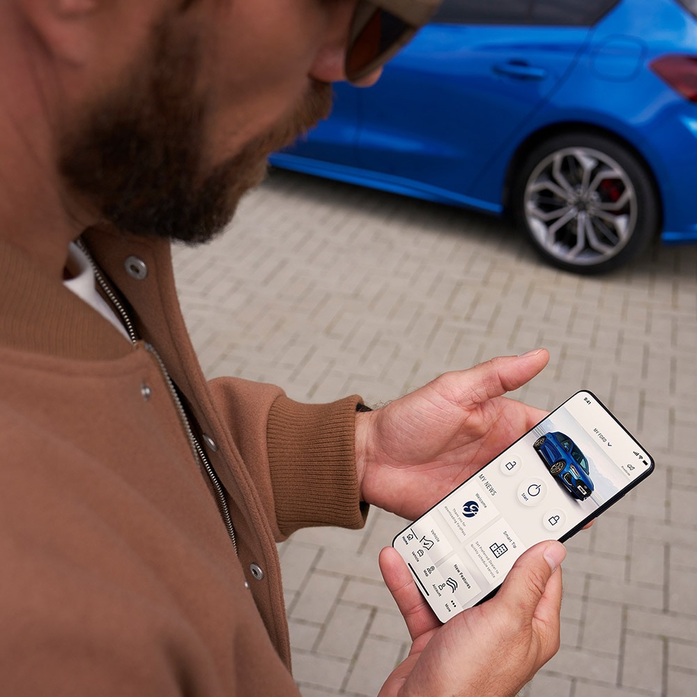 A person holding a smartphone displaying the FordPass™ app, with a blue Ford Focus in the background.
