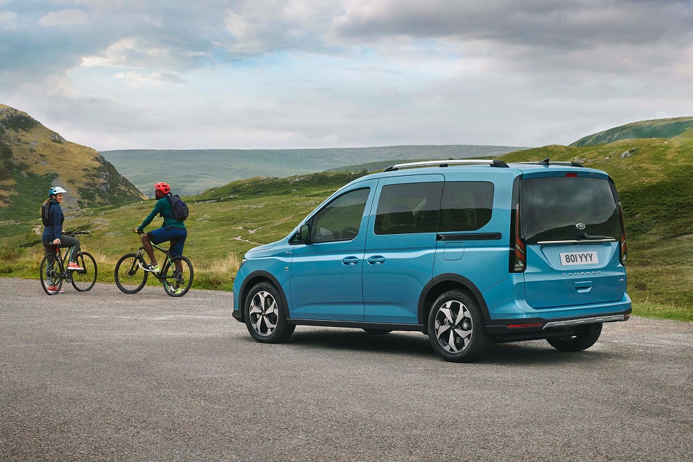 A blue Ford Tourneo Connect is parked on a road in a green, hilly landscape, with two people on bicycles nearby.