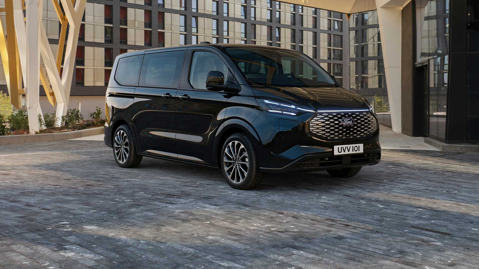 A black Ford E-Tourneo Custom is parked in front of a modern building.