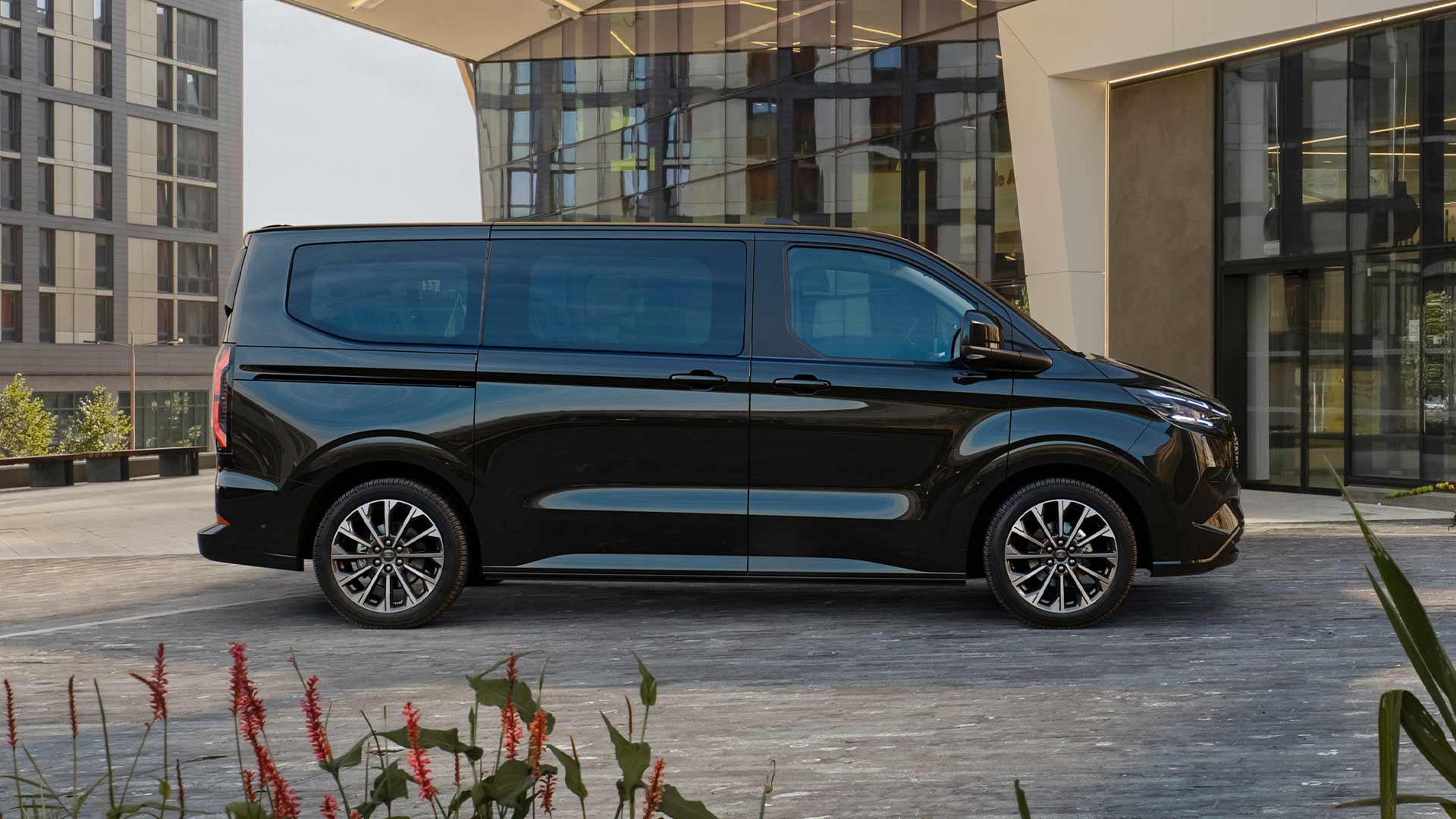 A black Ford E-Tourneo Custom is parked in front of a modern building.