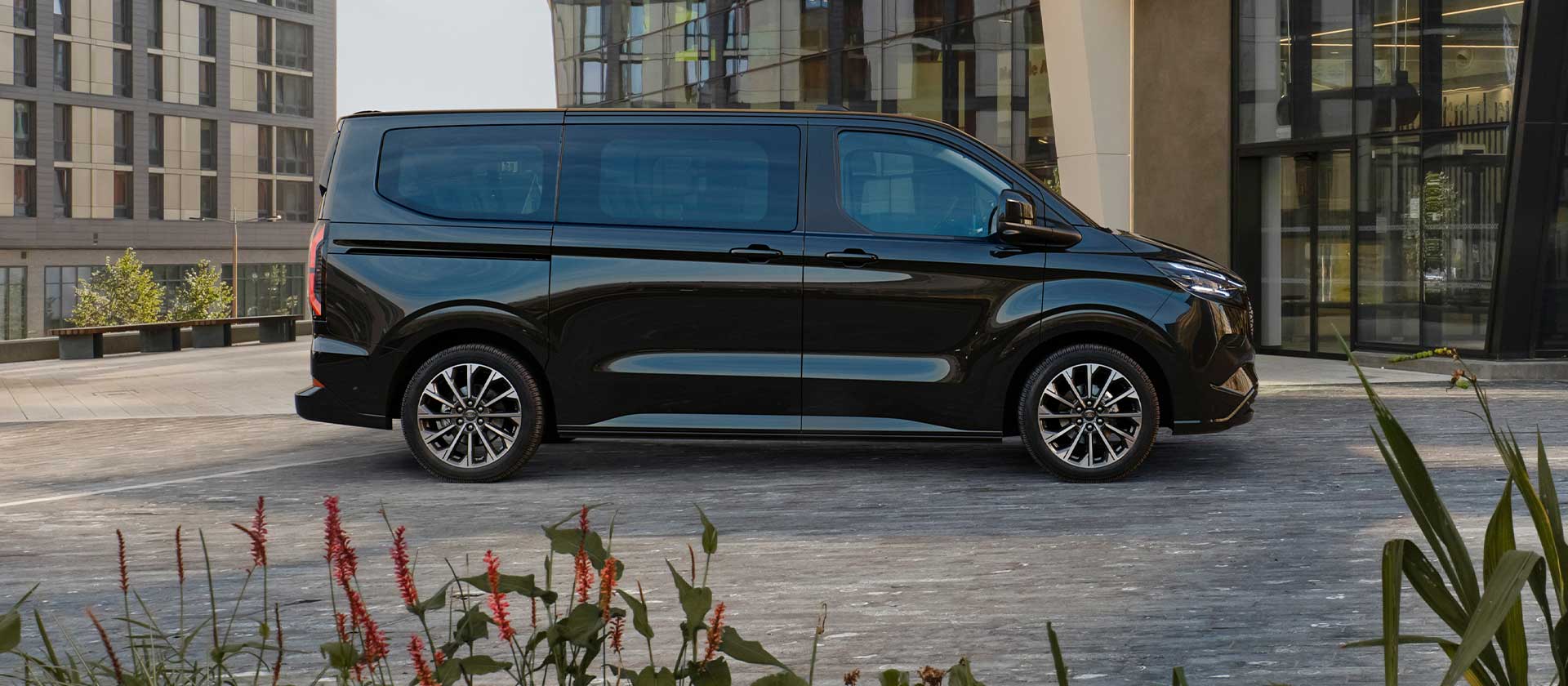 A black Ford E-Tourneo Custom is parked in front of a modern building.