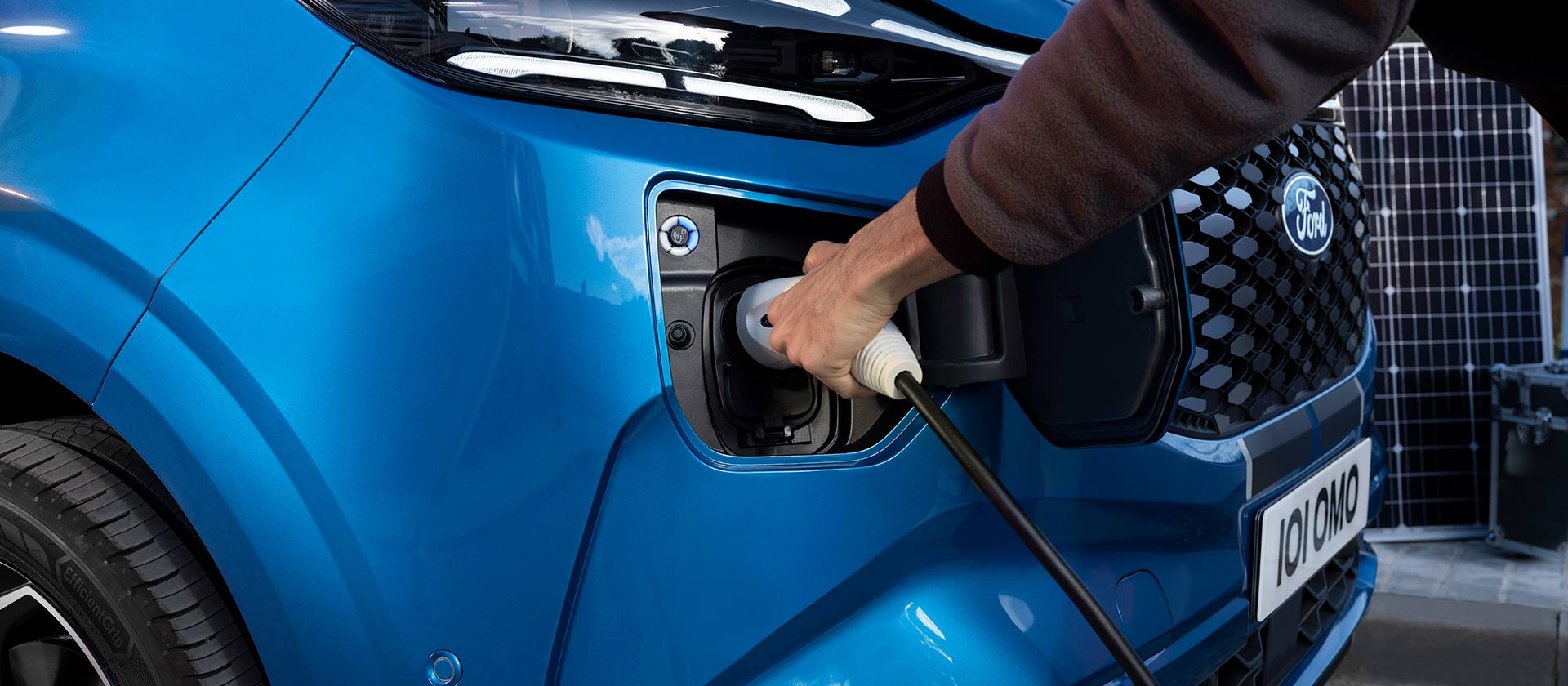 A hand inserts a charging cable into a blue Ford E-Tourneo Custom.