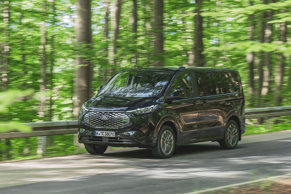 A black Ford E-Tourneo Custom drives along a tree-lined road.