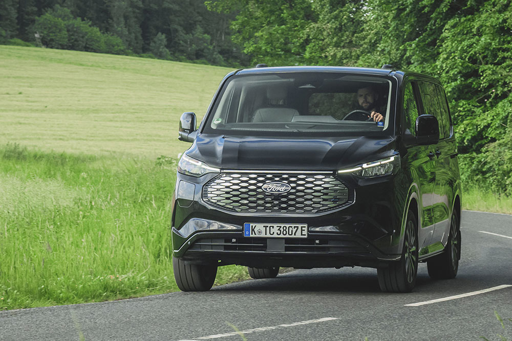 A black Ford E-Tourneo Custom drives on a countryside road.