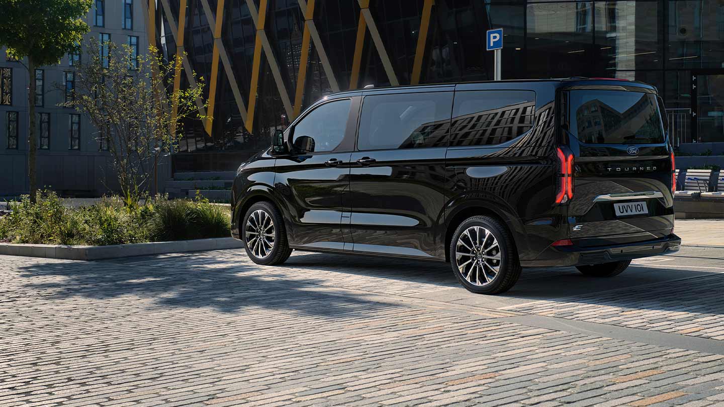 A black Ford E-Tourneo Custom is parked showing the side and rear in front of a modern building.