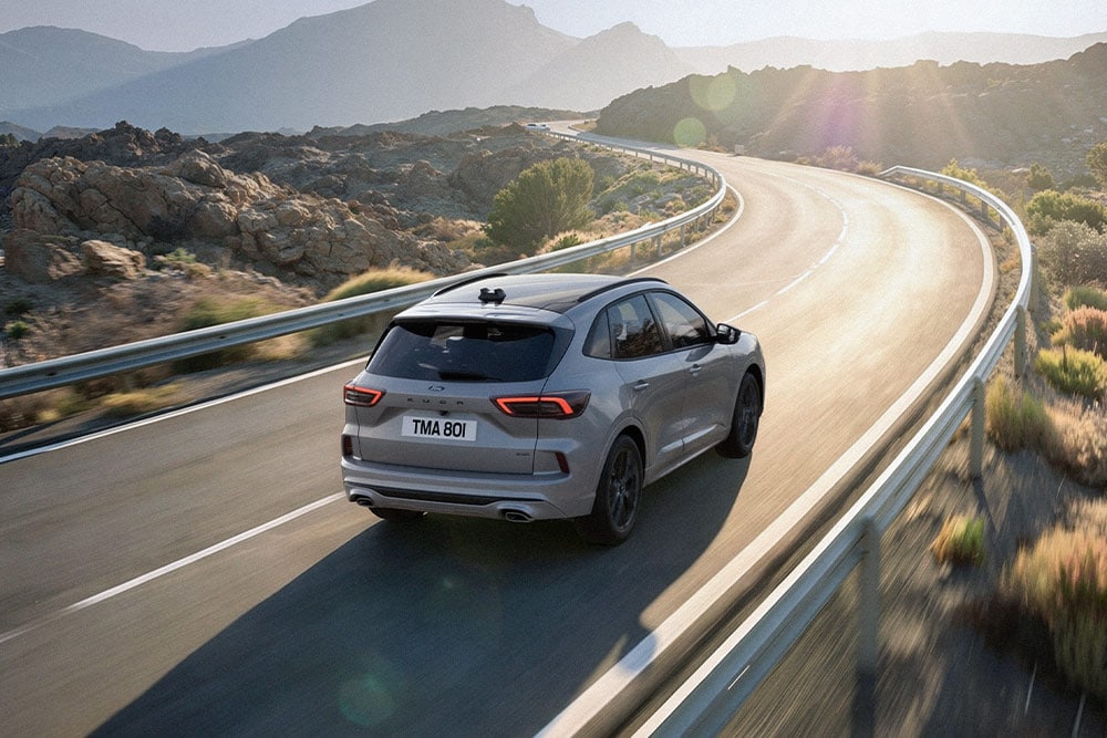 Silver Ford Kuga driving along a scenic route, sunny mountainous landscape in the background.