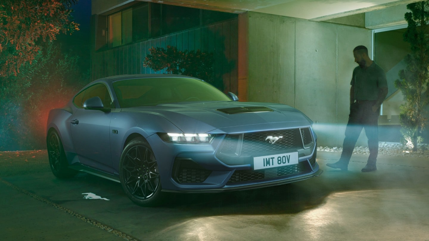 A blue Ford Mustang parked at night with its headlights on, a person standing nearby, and a Mustang logo projected on the ground.