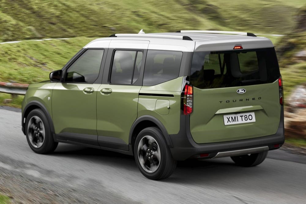 A green Ford Tourneo Courier is seen from the rear three-quarter angle, driving on a winding road with green hills.