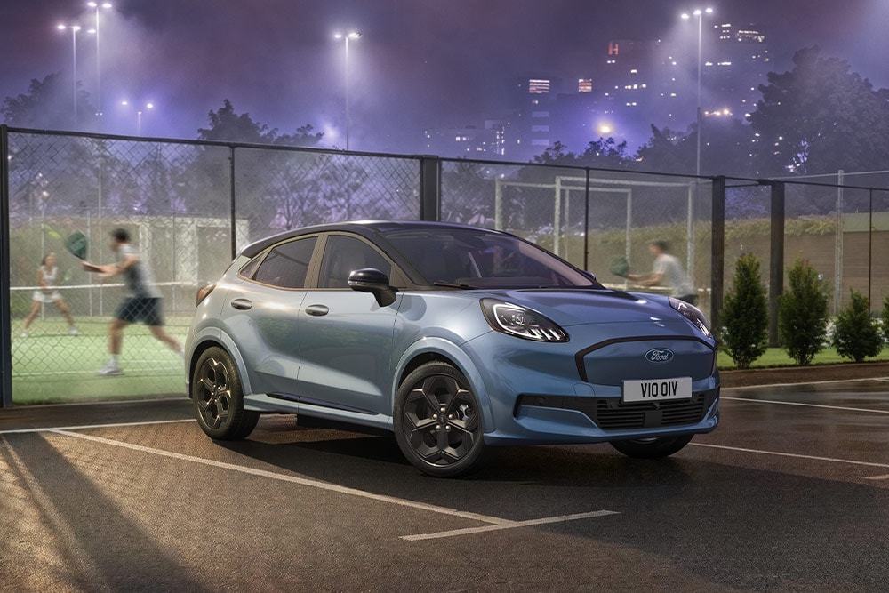 A blue Ford Puma Gen-E parked next to a tennis court, with city lights in the background.