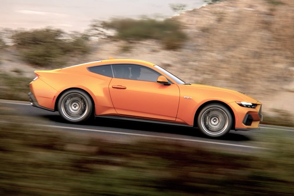 A side view of the Ford Mustang GT Fastback in Orange Fury paint, driving along a road.