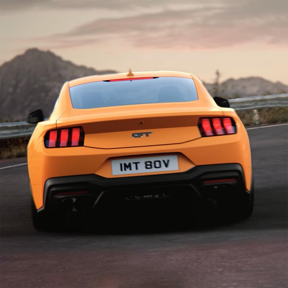 The rear of a Ford Mustang GT Fastback in Orange Fury paint, driving a tight curve in a country road.