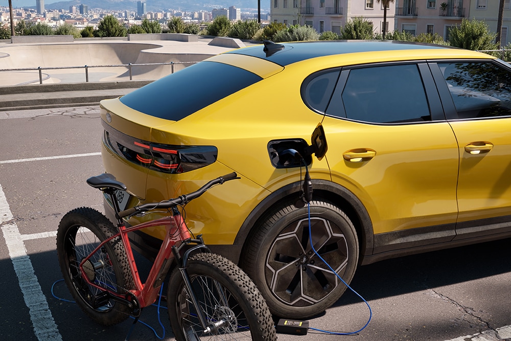 Yellow Ford Capri® parked, charging a red electric bicycle in an urban cityscape.