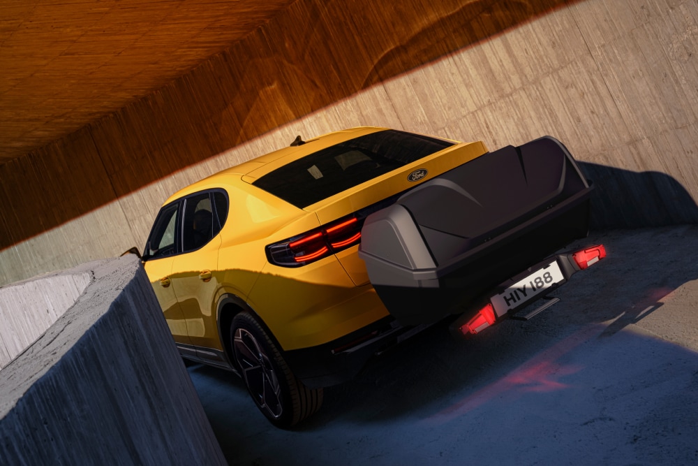 Yellow Ford Capri parked in a modern concrete structure, rear view showing a luggage box attached to the rear with a luggage box carrier.