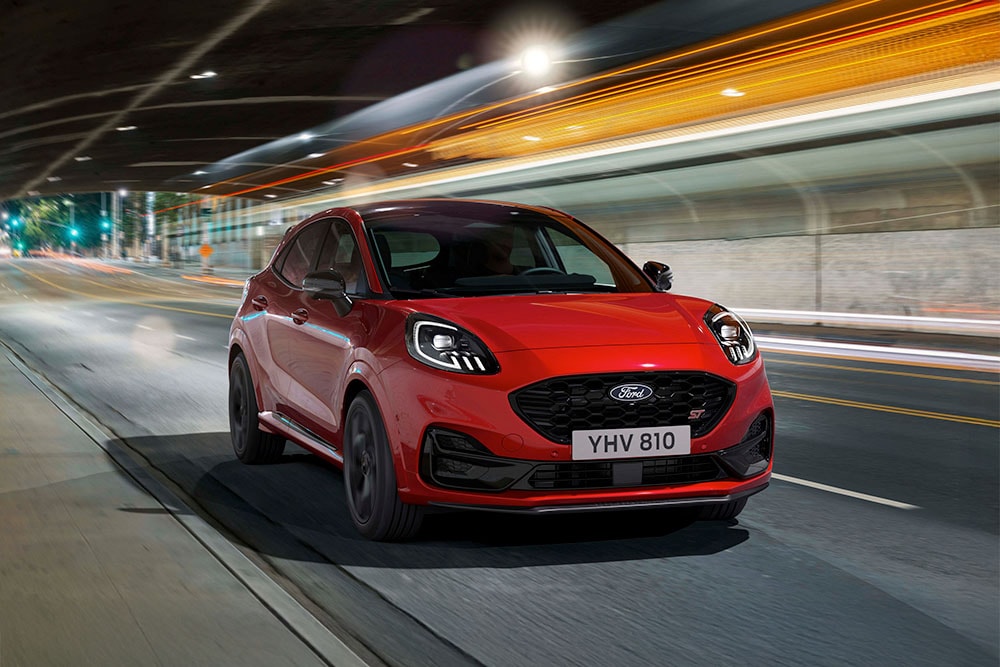 A red Ford Puma ST sporty SUV driving along a brightly lit city road at night.
