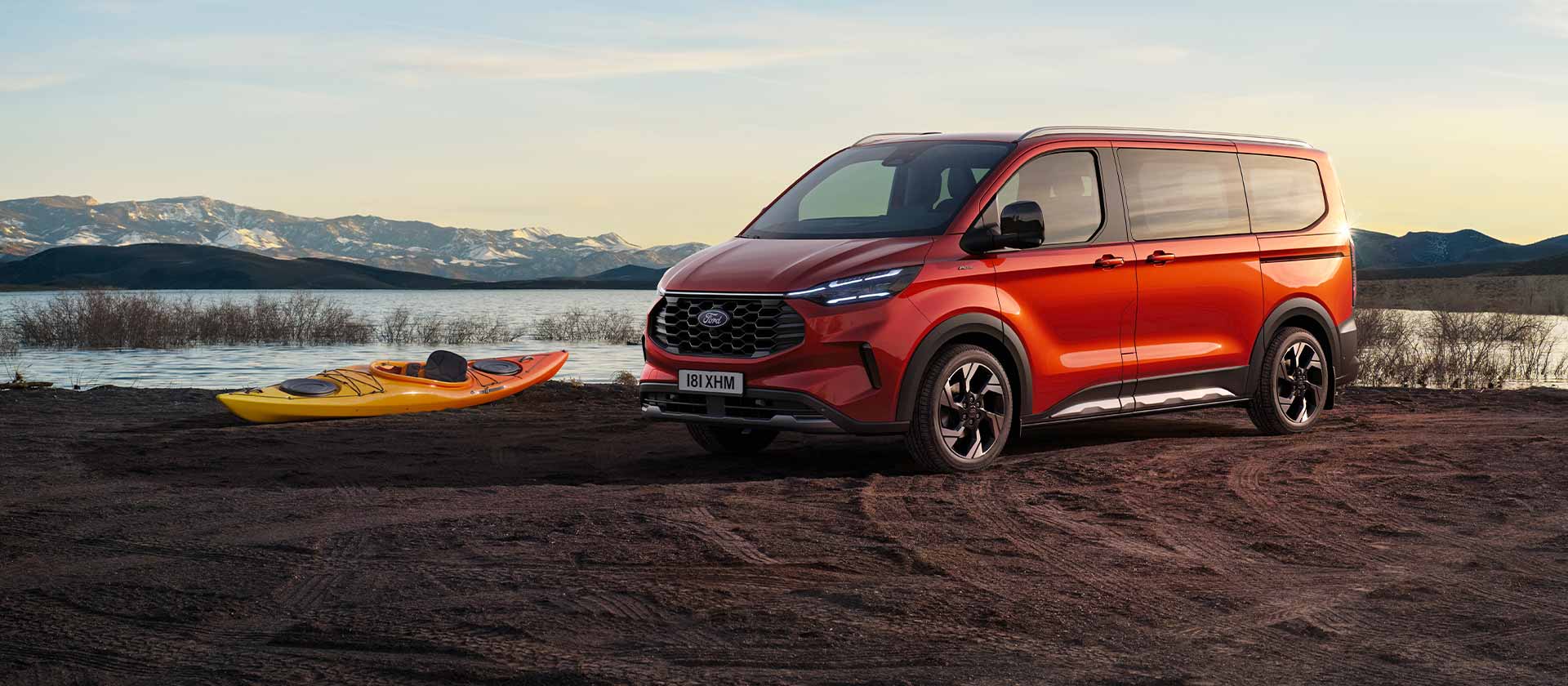A red Ford Tourneo Custom van is parked near a lake with a kayak. 