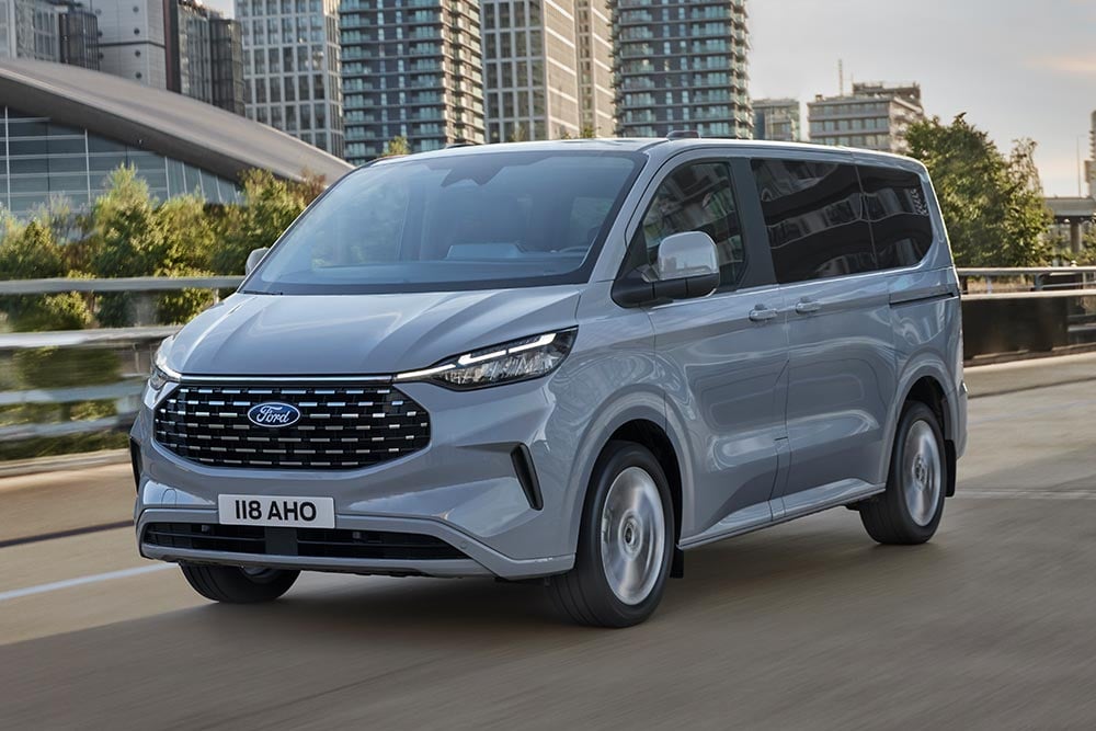 A silver Ford Tourneo Custom drives on a city road.