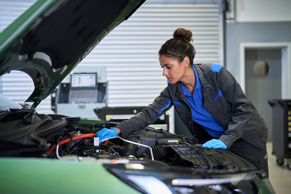 Technician in blue gloves working on the engine of a green car with the bonnet open.