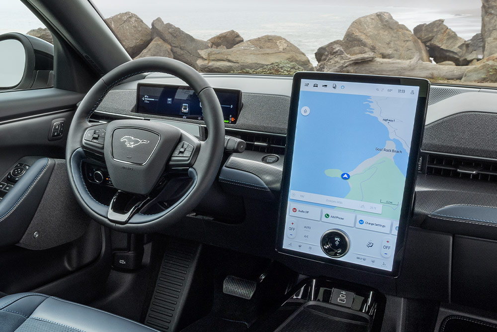 Interior of a Ford Mustang Mach-E, showing the dashboard, steering wheel, and large touchscreen with a map, parked by a rocky coast.