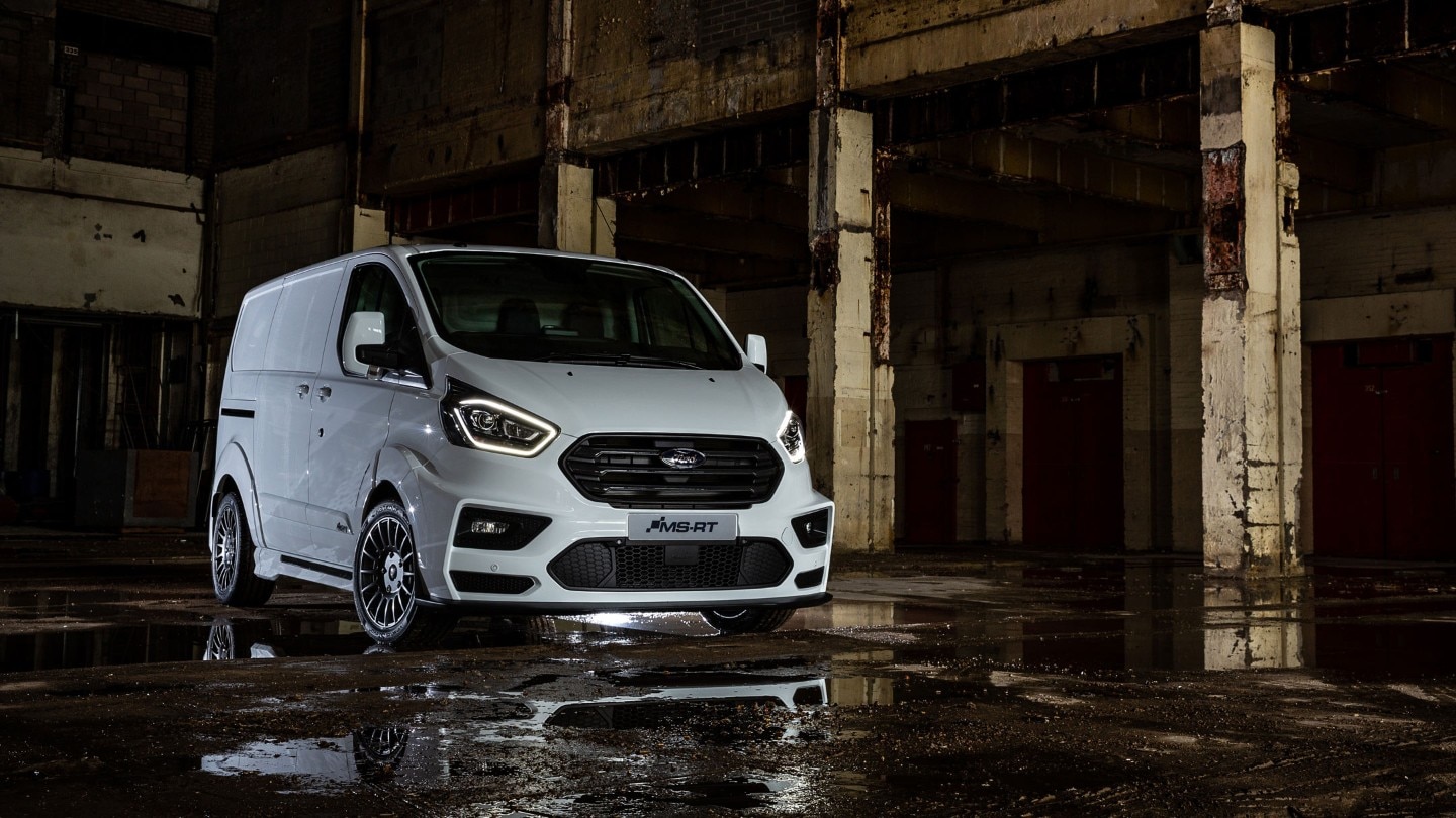 Ford Transit Custom MS-RT 3-quarter view, parked on a cobbled side street against a residential building backdrop. 