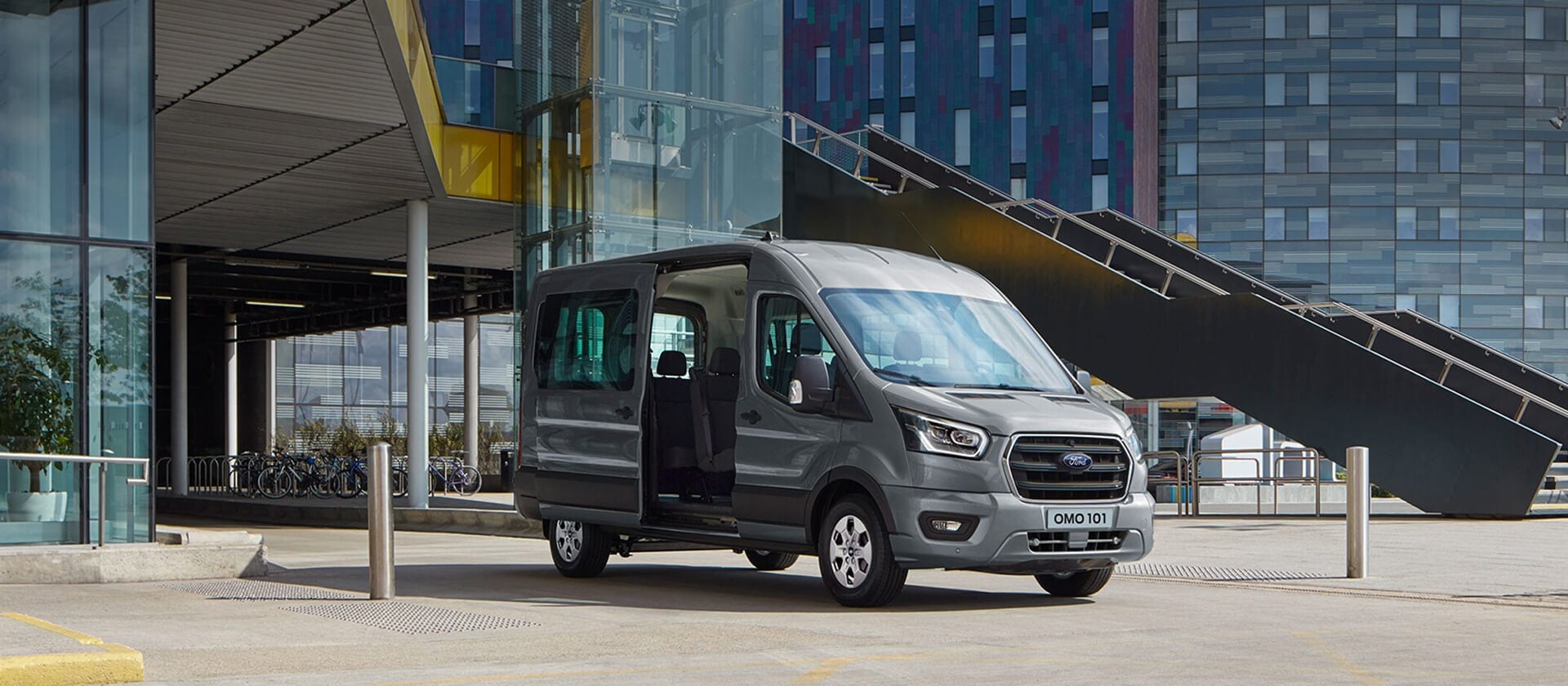 Grey Ford Transit Minibus parked in front of a modern building, with its side door open revealing the interior.