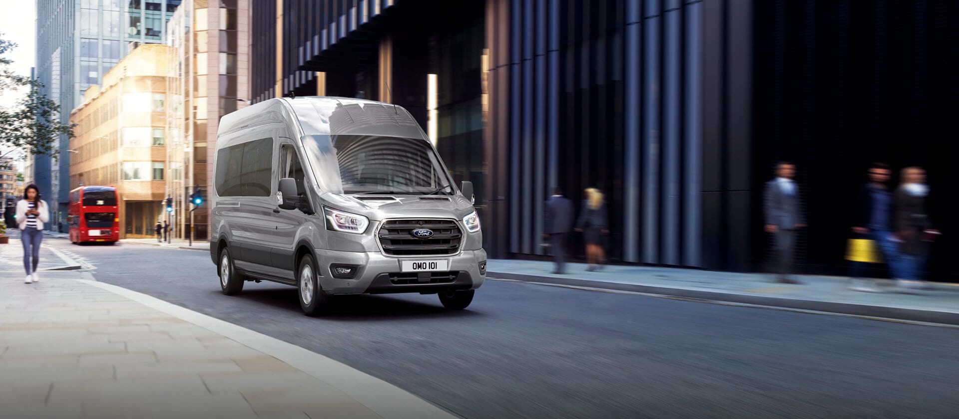 Silver Ford Transit Minibus driving on a city street with a red double-decker bus and people in the background.