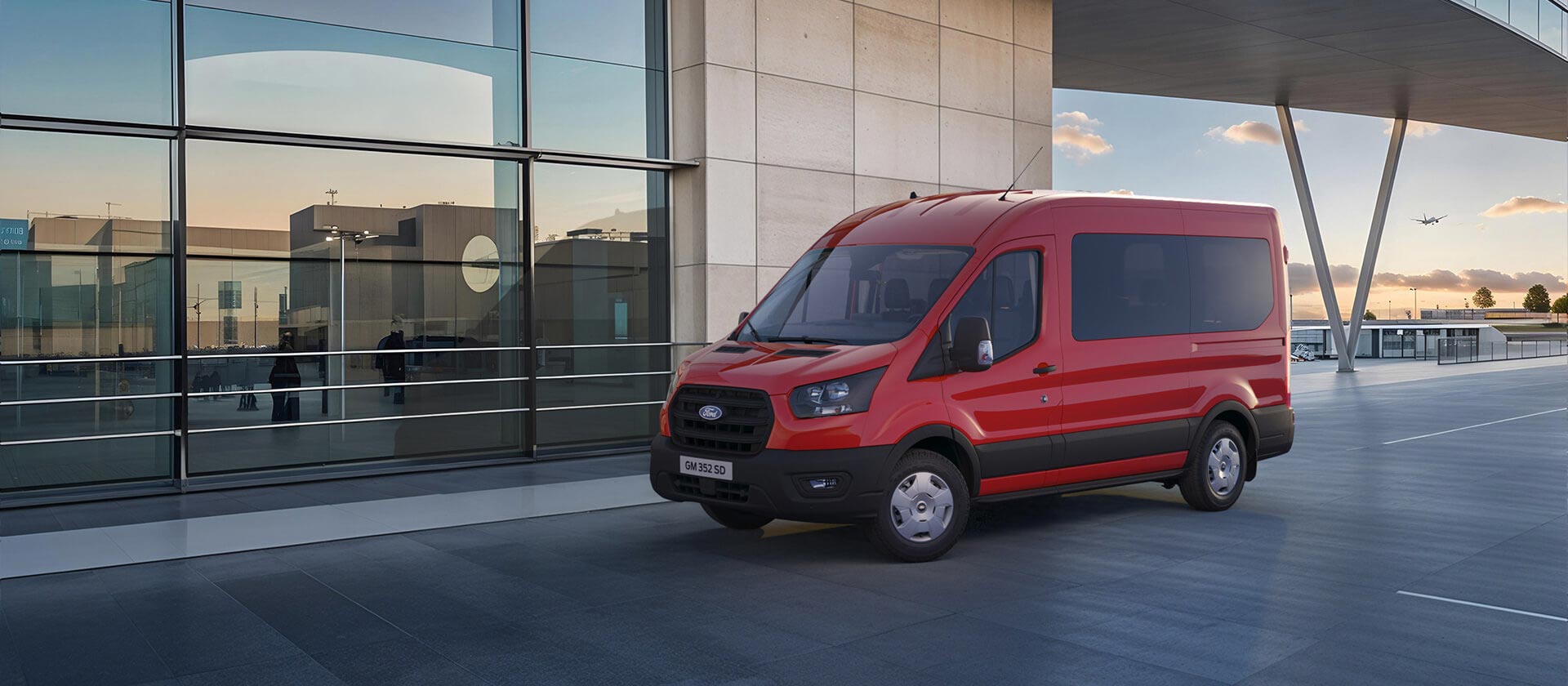 Front 3/4 view of a red Ford Transit Minibus parked outside a modern airport terminal with a plane in the distance.