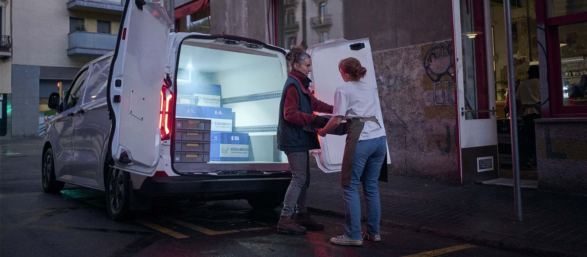 A Ford delivery van with open doors is parked on a dimly lit street. Two women are unloading packages.
