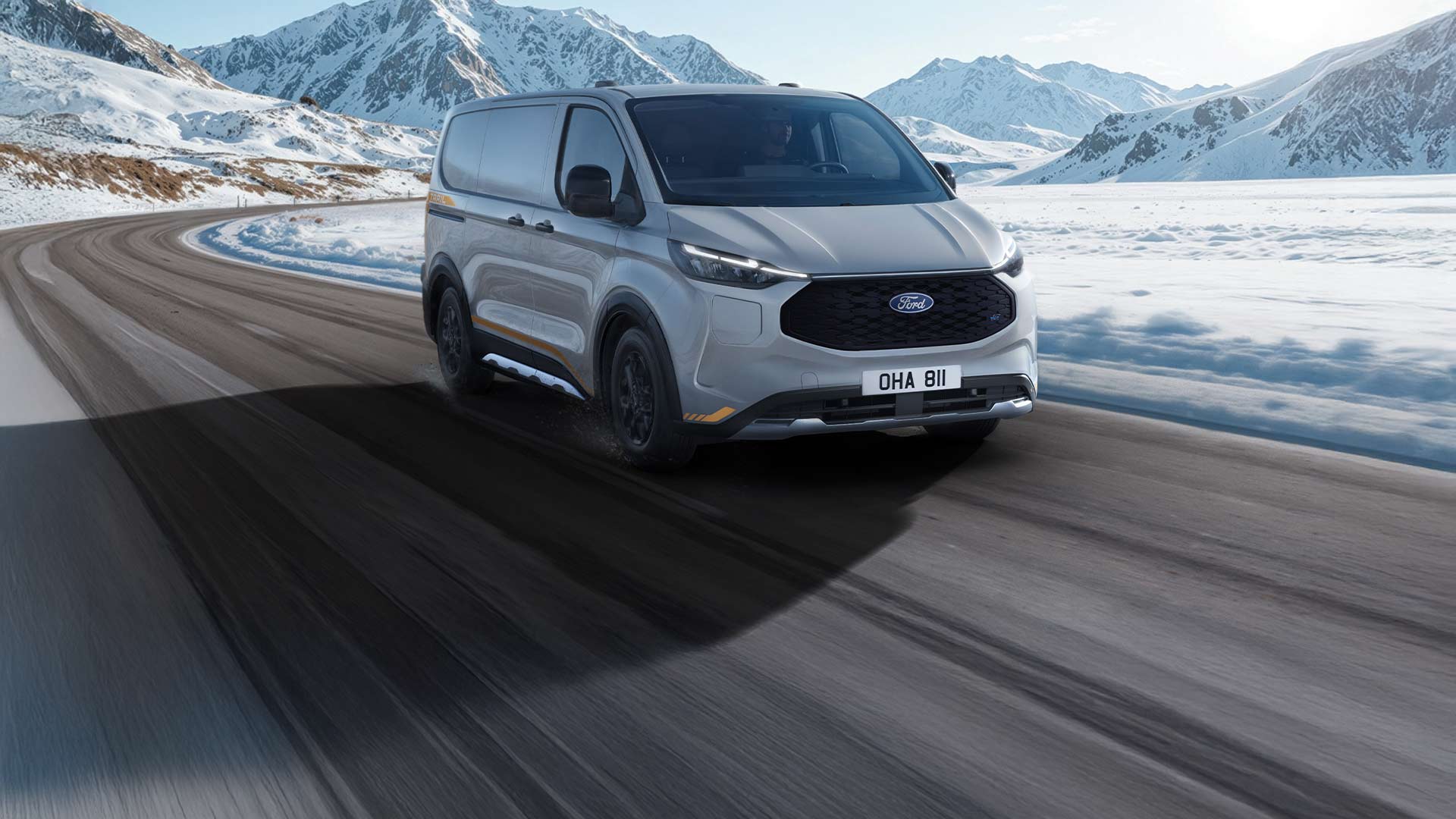 A silver Ford E-Transit Custom driving on a snowy mountain road under a clear blue sky.