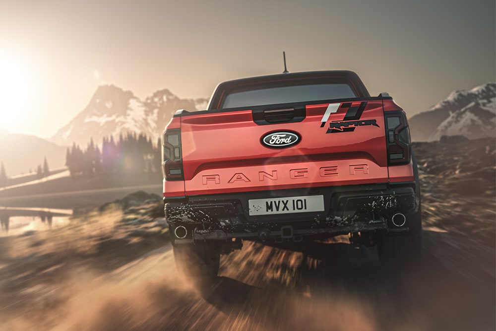 Rear view of a red Ford Ranger Raptor driving on a dirt road with mountains in the background.