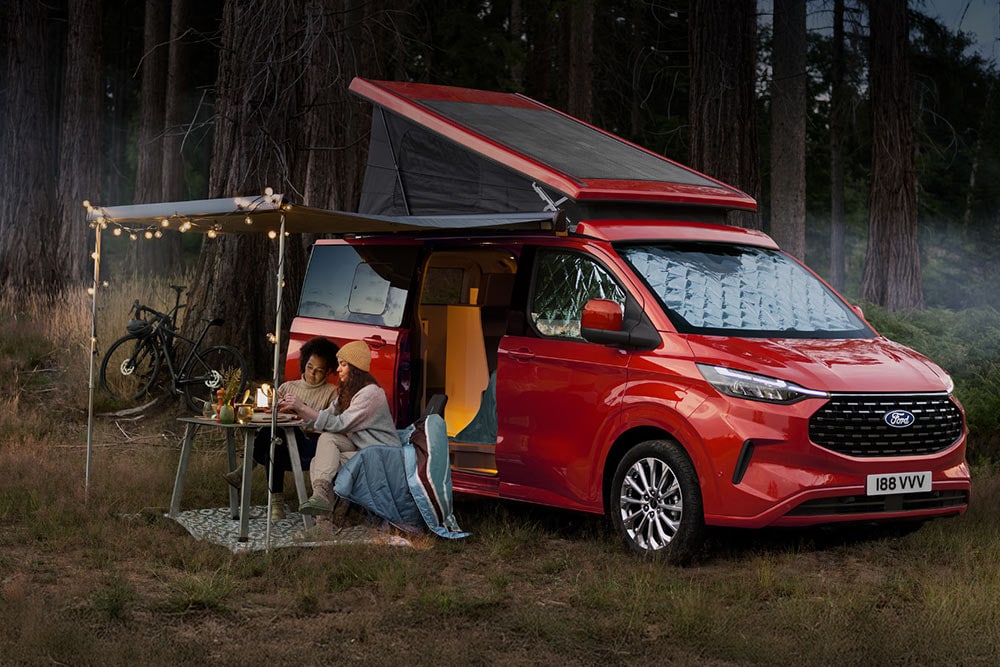 Two women sit under an awning from the open side doors of a red Ford Transit Custom Nugget, with the tilt roof up.