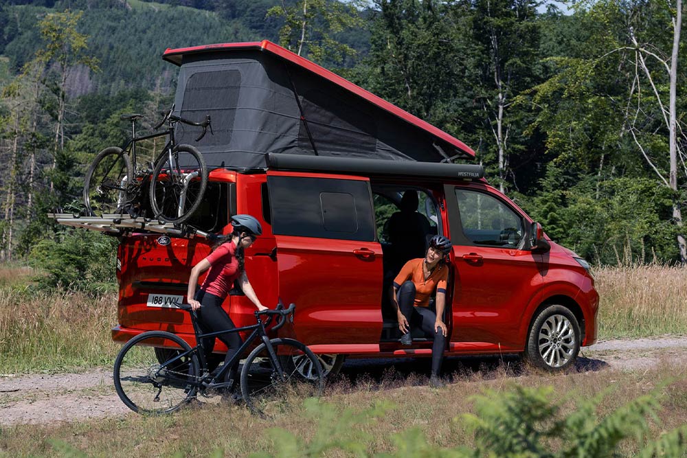 People unload bikes from the bike rack of a red Ford Transit Custom Nugget.