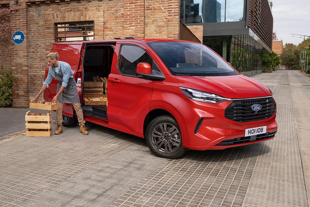 Delivery driver loading packages into the side door of a red Ford Transit Custom.