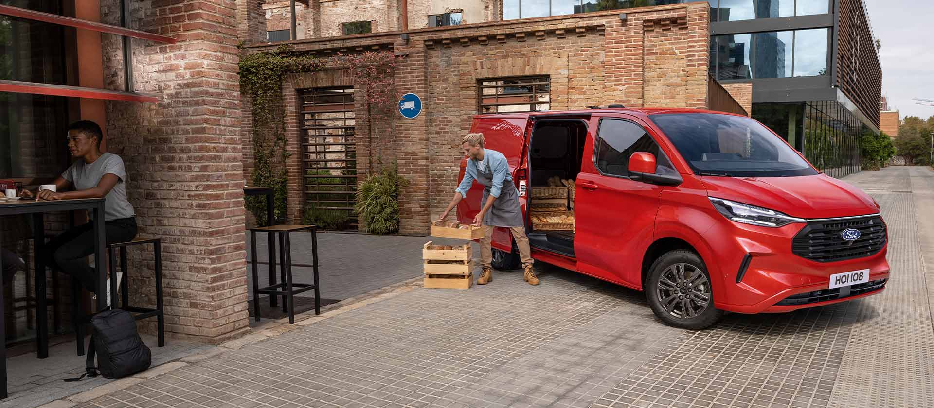 A person unloads crates from a red Ford Transit Custom van parked in front of a coffee shop. 