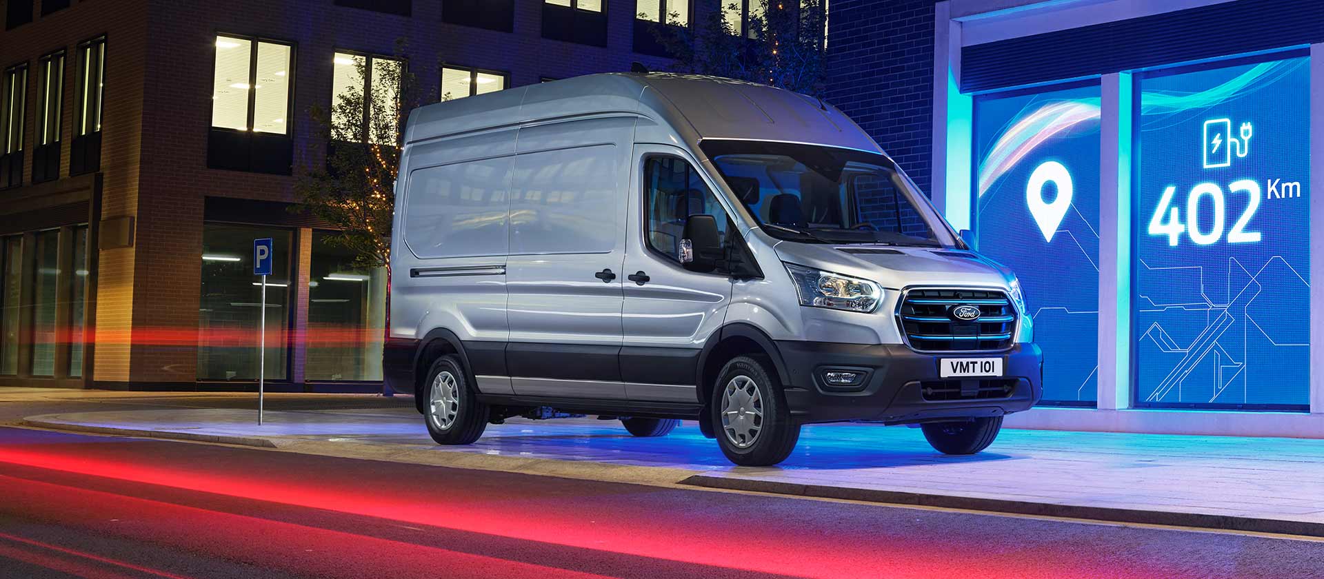 Silver Ford E-Transit electric van parked on a city street at night with range displayed.