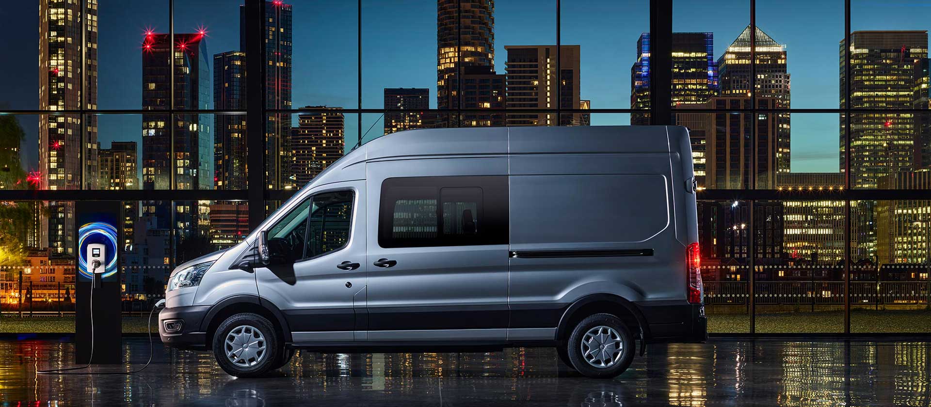 Silver Ford E-Transit Double Cab-in-Van charging with a city skyline background.
